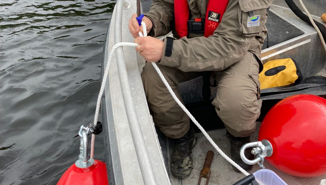 Person i båt gör iordning en boj, med en skylt. Foto.