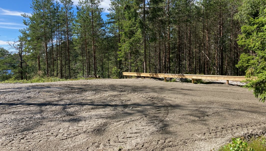 Grusad parkering med ett träräcke mot skog. Foto.