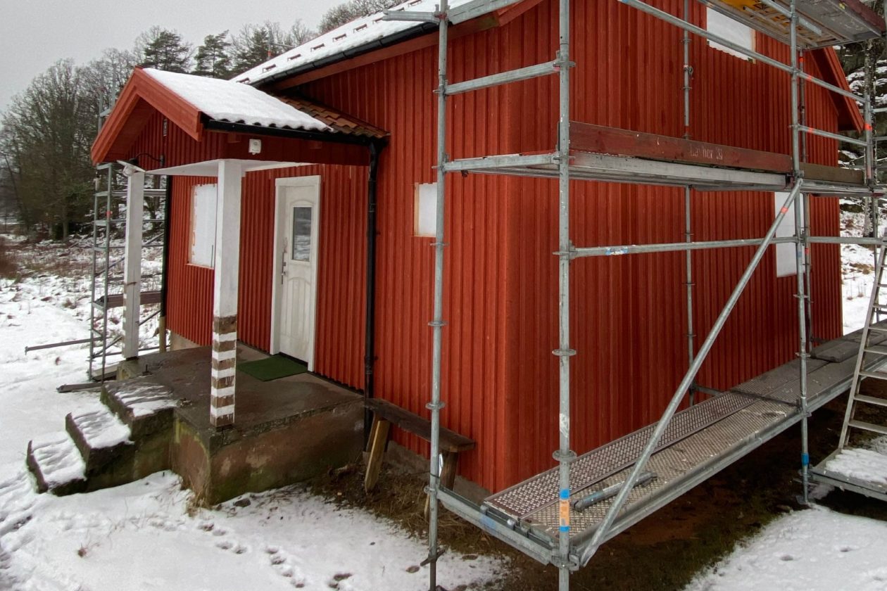En röd stuga med en byggställning på ena sidan. Foto med texten "Västkuststiftelsen - vi söker en aktör till Bottenstugan"..
