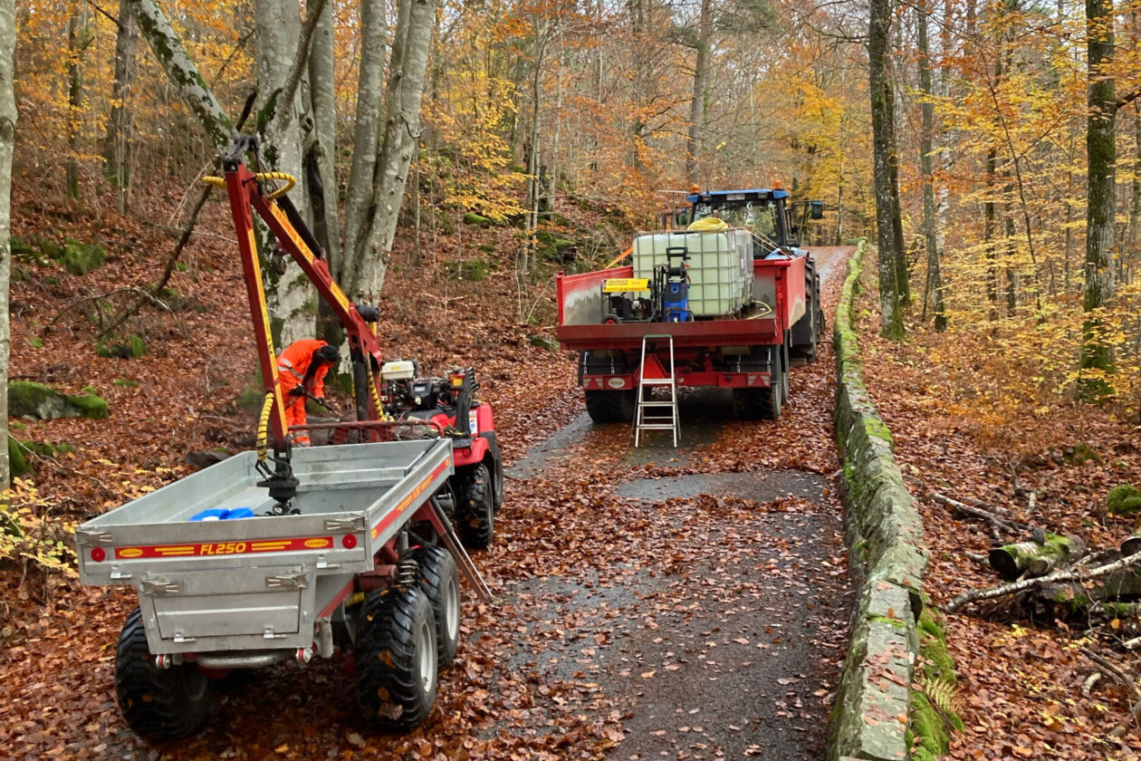 En traktor och en fyrhjuling med släp vardera, som blockerar en väg med en stenmur längs ena sidan. Foto.