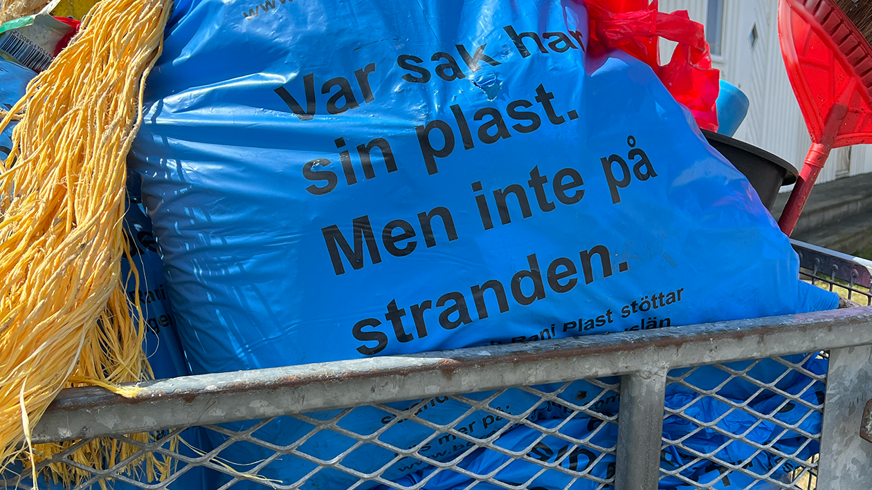 Blå sopsäck med strandskräp som ligger i en vagn med annat skräp. På sopsäcken står texten "Var sak har sin plast, men inte på stranden".