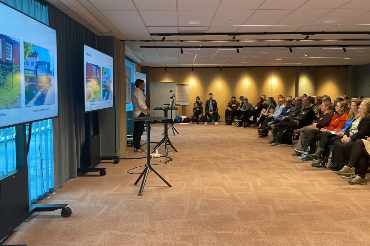 Stor grupp personer sitter och ser på en talare med flea skärmar bakom sig. Foto.