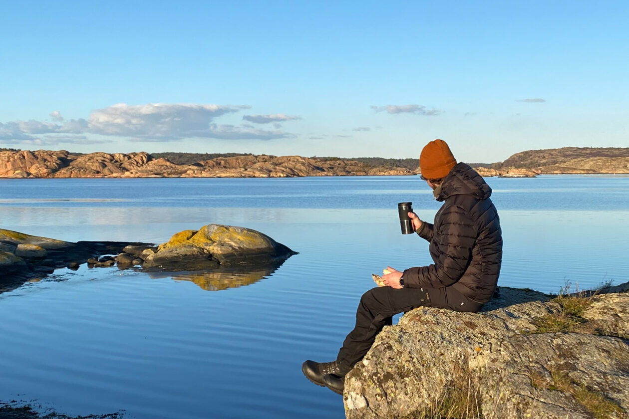 Person sitter med fika påå en klippa vid havet. Foto.