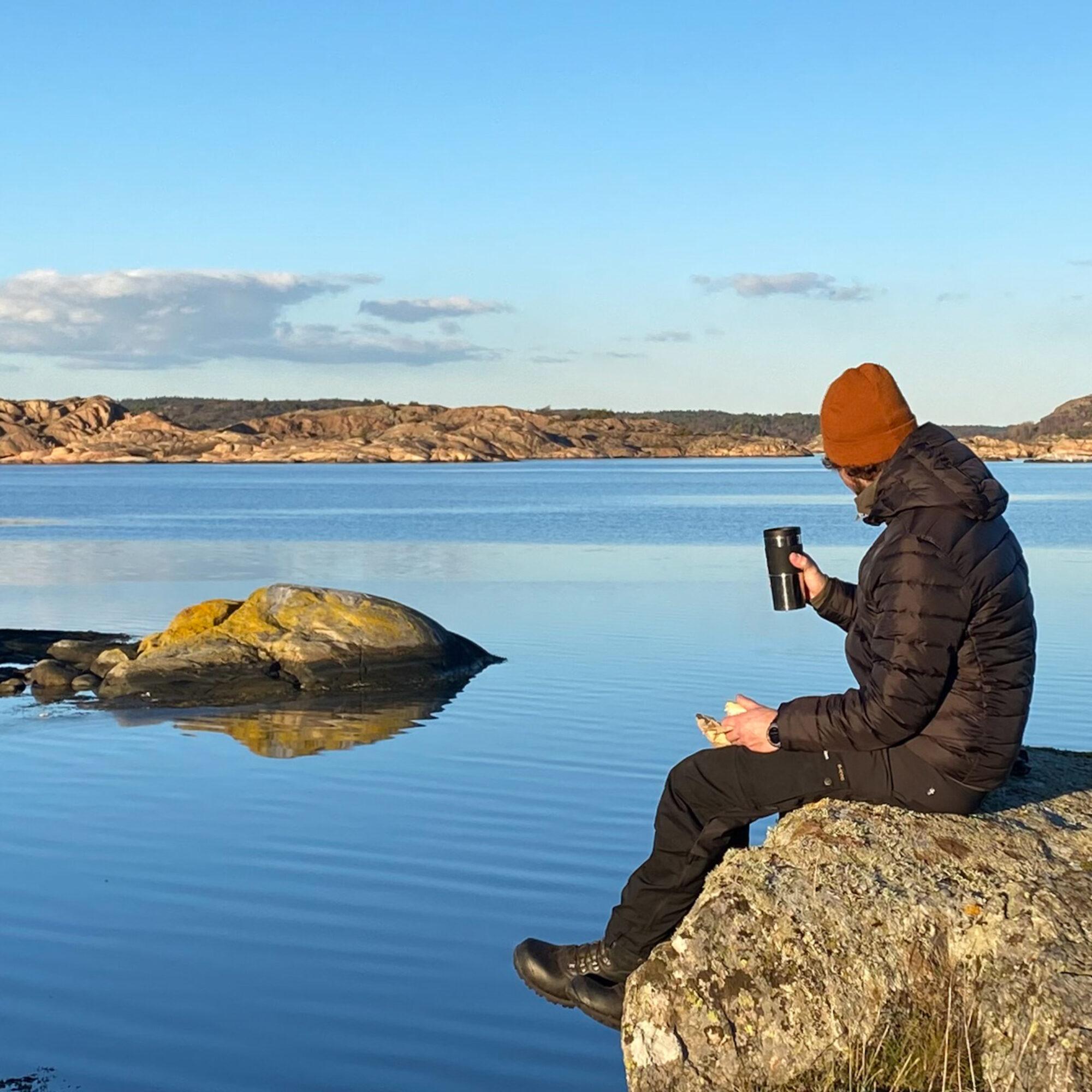 Person sitter med fika påå en klippa vid havet. Foto.