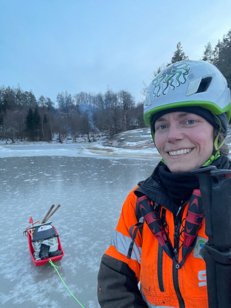 Närbild på person står på isen och en pulka med gasoltub och annan utrustning och ännu längre bort ser man rök från en rishög som eldas. Foto.