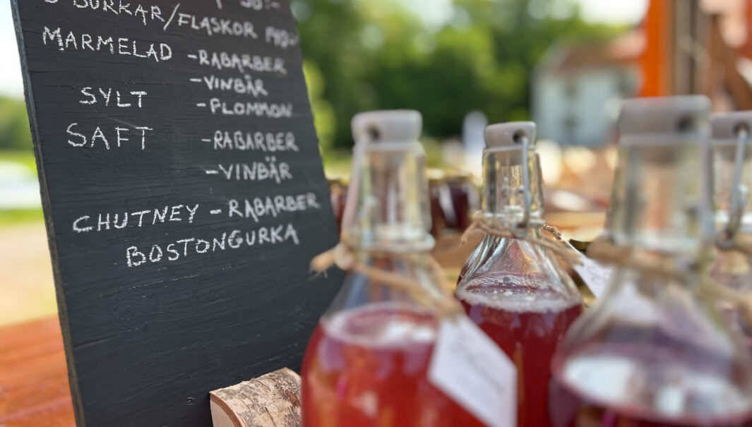 Glasflaskor med röd saft i förgrunden och i bakgrunden en tavla med priser på produkter. Foto.