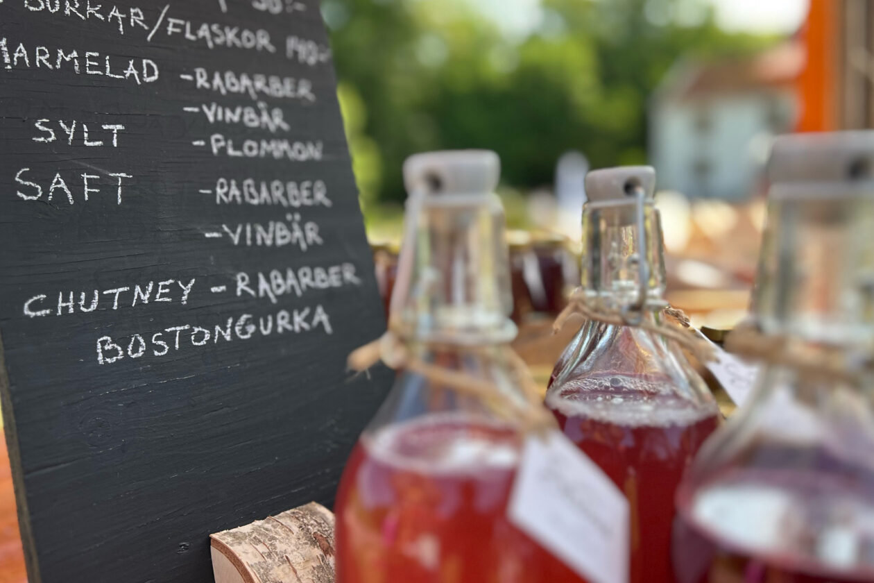 Glasflaskor med röd saft i förgrunden och i bakgrunden en tavla med priser på produkter. Foto.