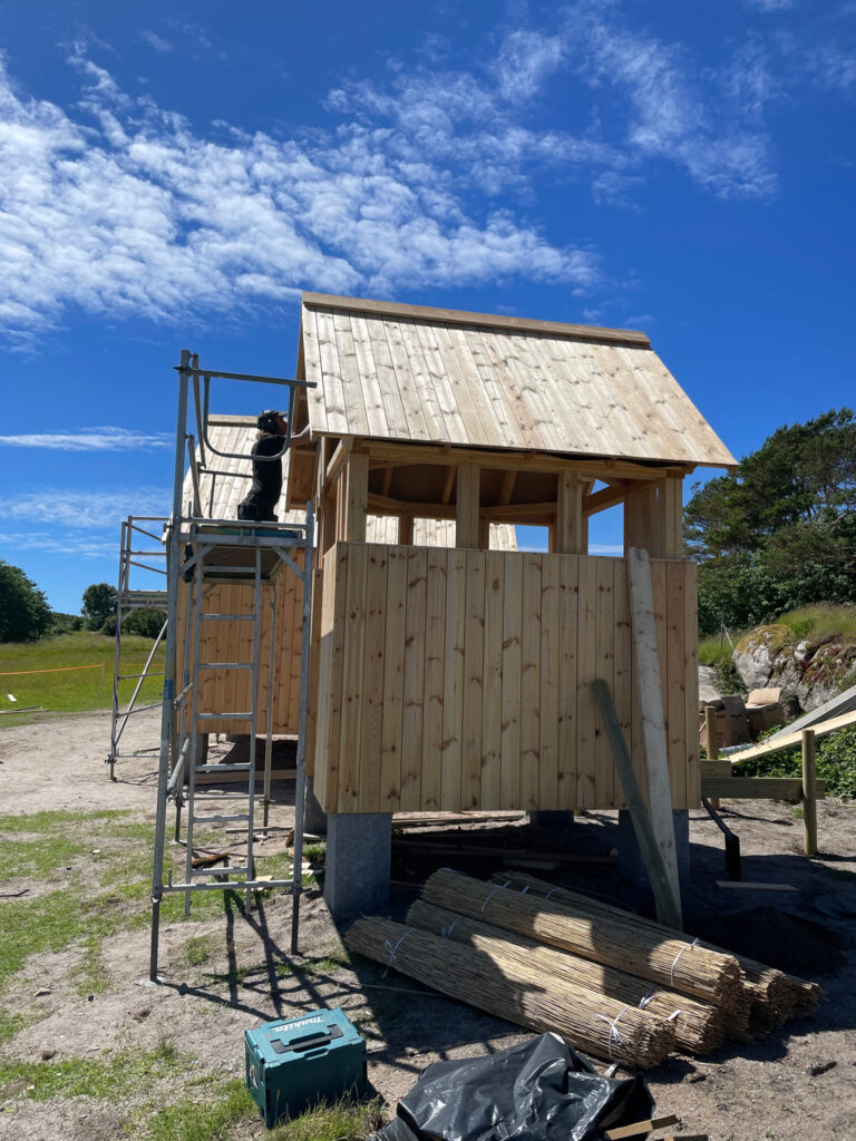 en snickare arbetar från en byggställning vid en liten träbyggnad. Foto.
