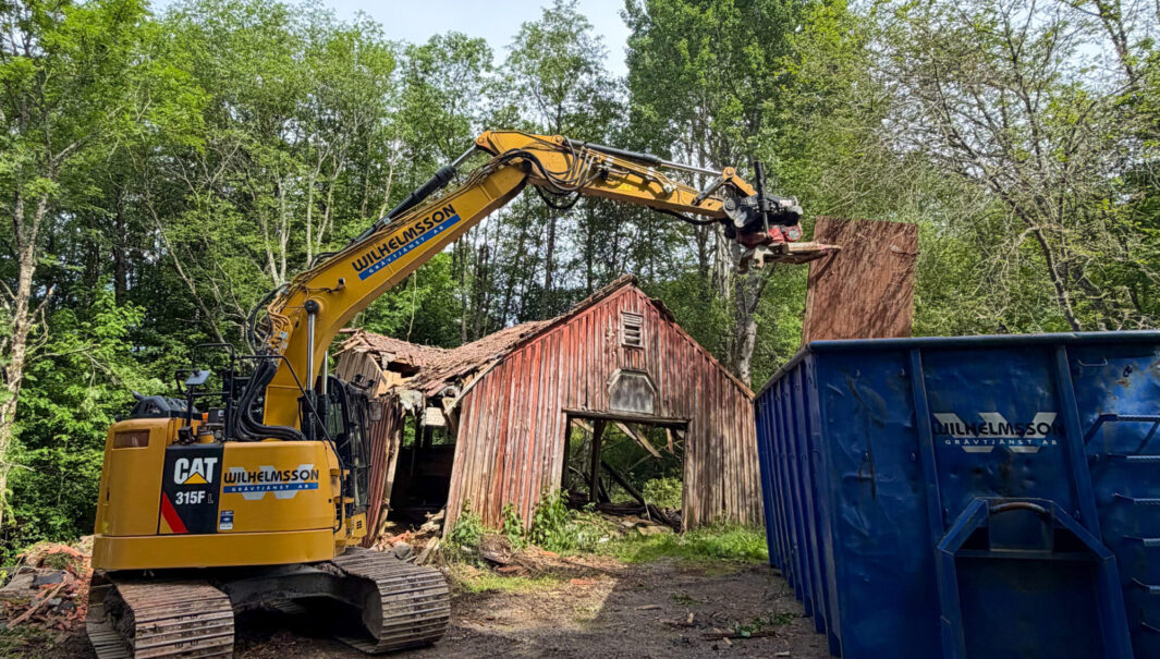 En grävmaskin och en blå container framför en fallfärdig röd lada. Foto.