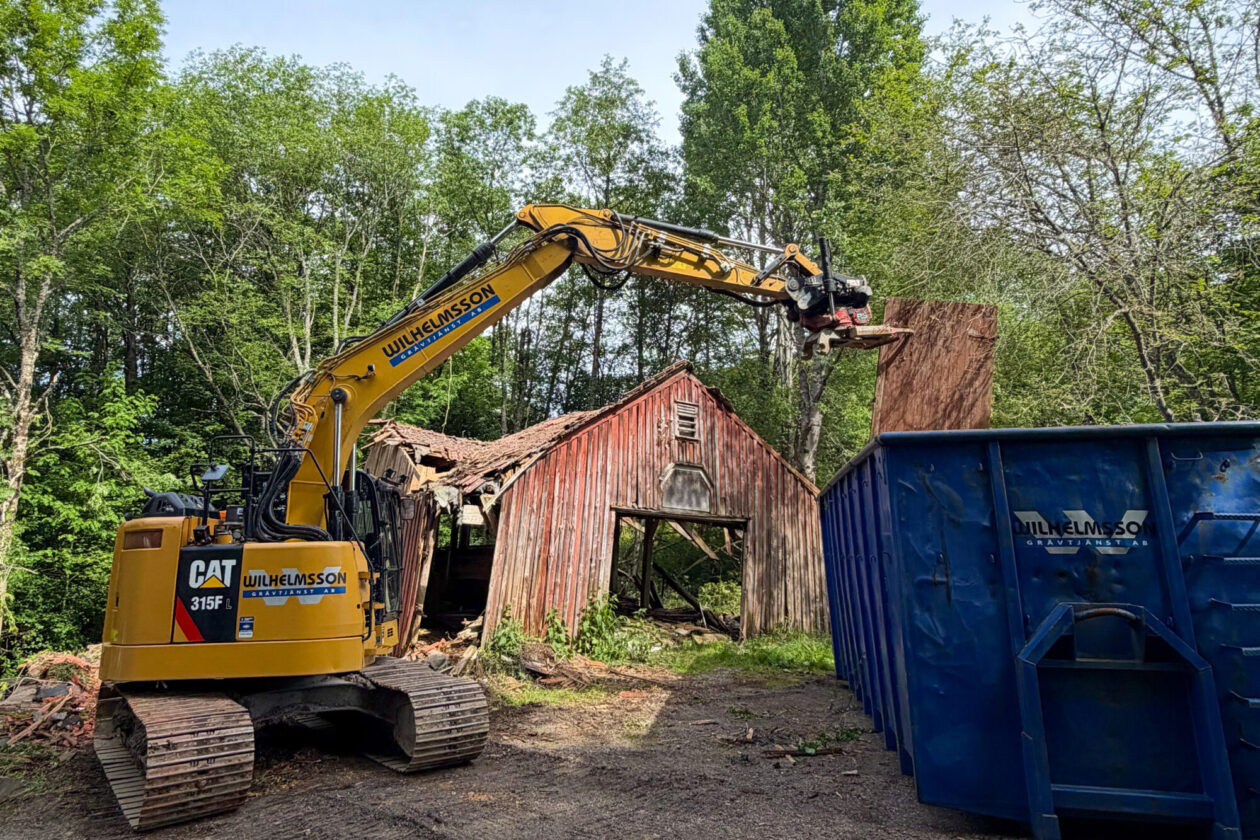En grävmaskin och en blå container framför en fallfärdig röd lada. Foto.