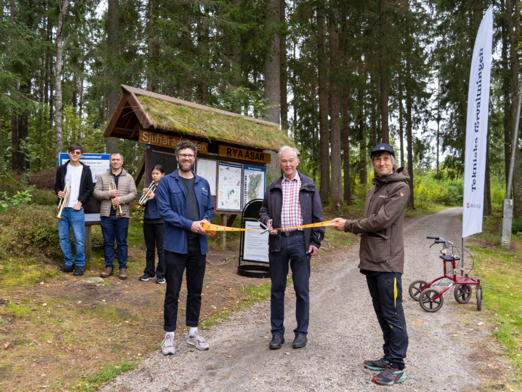 Tre personer på rad. Personen i mitten klipper i ett band som de andra två håller. I bakgrunden står tre personer med var sin trumpet. Foto.