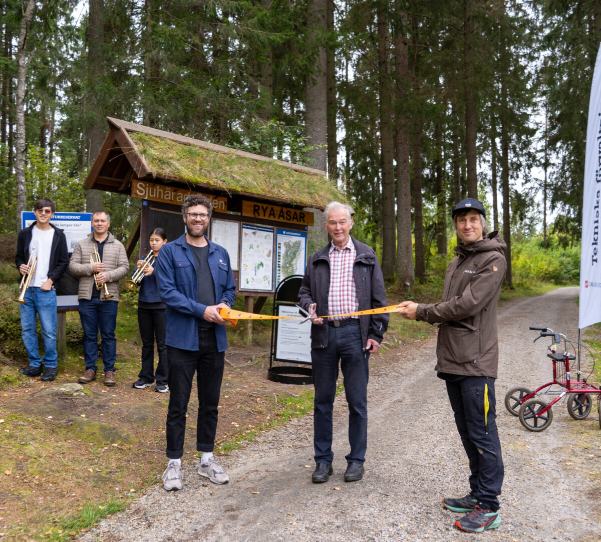 Tre personer på rad. Personen i mitten klipper i ett band som de andra två håller. I bakgrunden står tre personer med var sin trumpet. Foto.