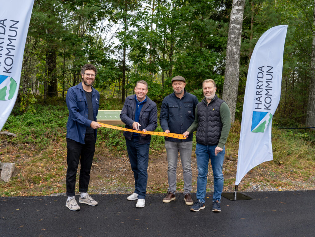 Fyra personer på rad omgivna av beachflaggor. Två av personerna håller i ett orange band medan en trejde klipper. Foto.