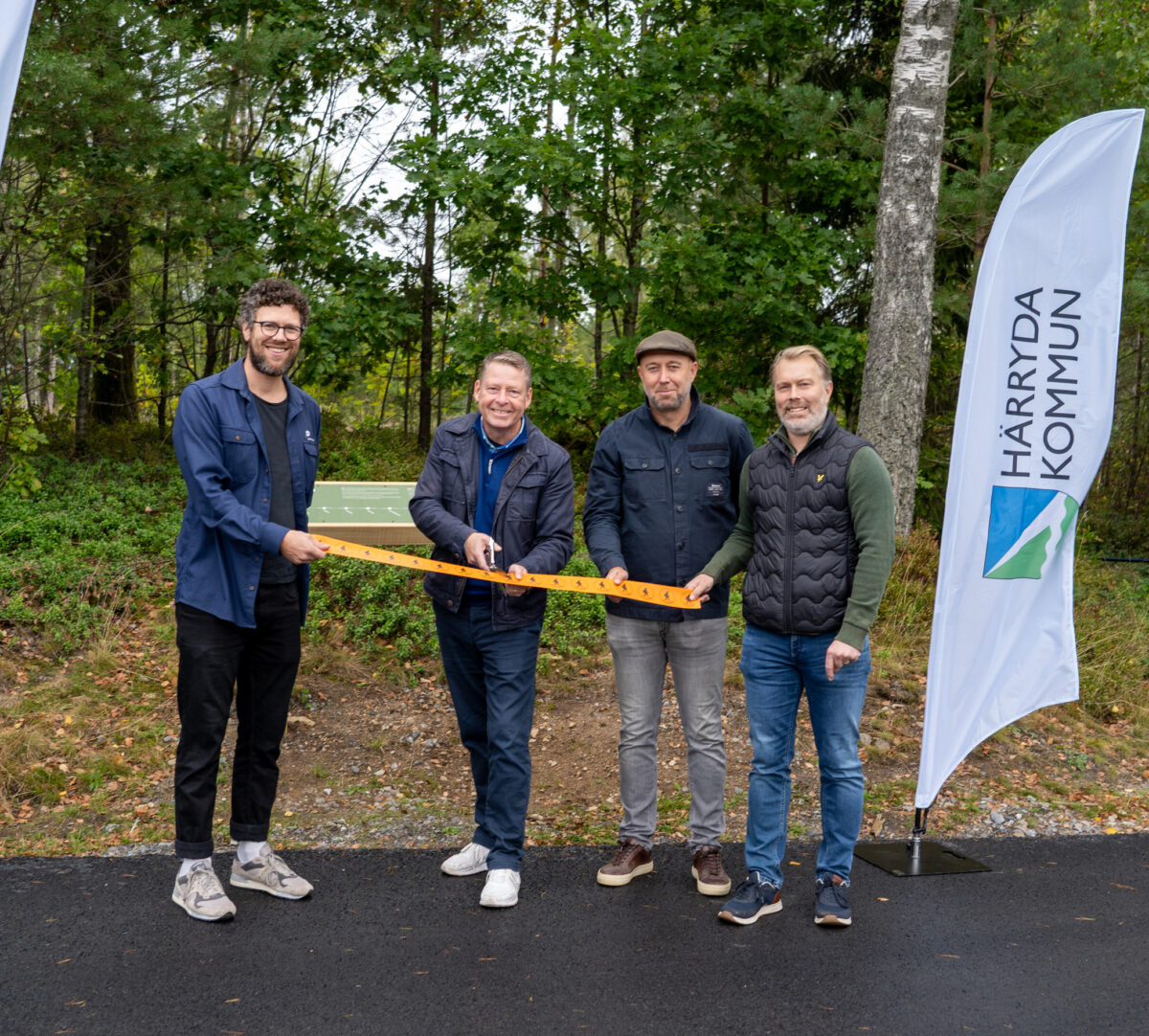 Fyra personer på rad omgivna av beachflaggor. Två av personerna håller i ett orange band medan en trejde klipper. Foto.