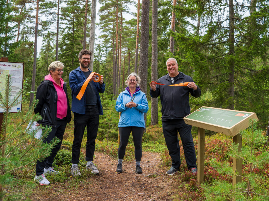 Fyra skrattande personer på rad i skogen vid en etappskylt. Två håller i ett avklippt orange band och en håller i en sax. Foto.