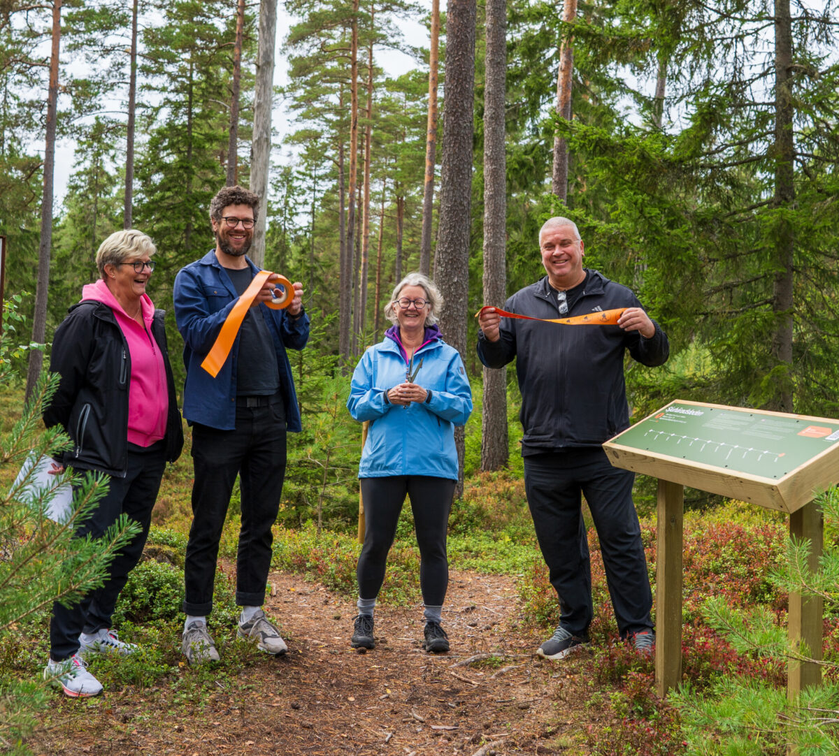 Fyra skrattande personer på rad i skogen vid en etappskylt. Två håller i ett avklippt orange band och en håller i en sax. Foto.