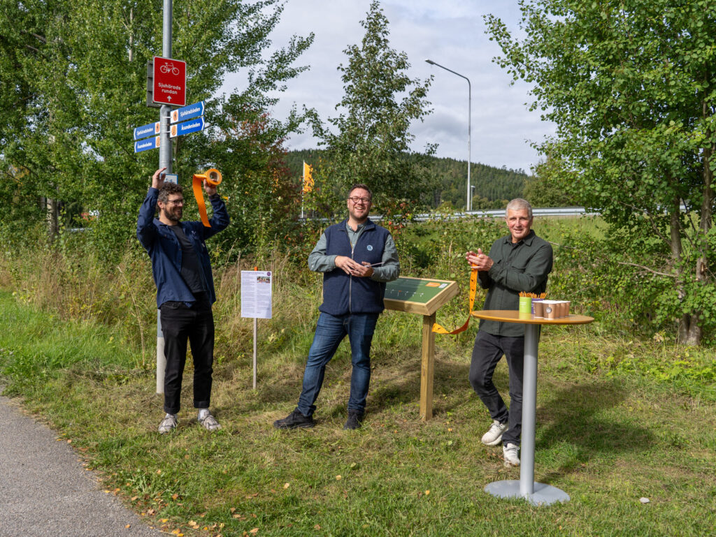 Tre glada personer på rad. Personen till vänster firar med händerna uppe, personen i mitten håller i en sax och personen till höger håller i en bit orange band. Foto.