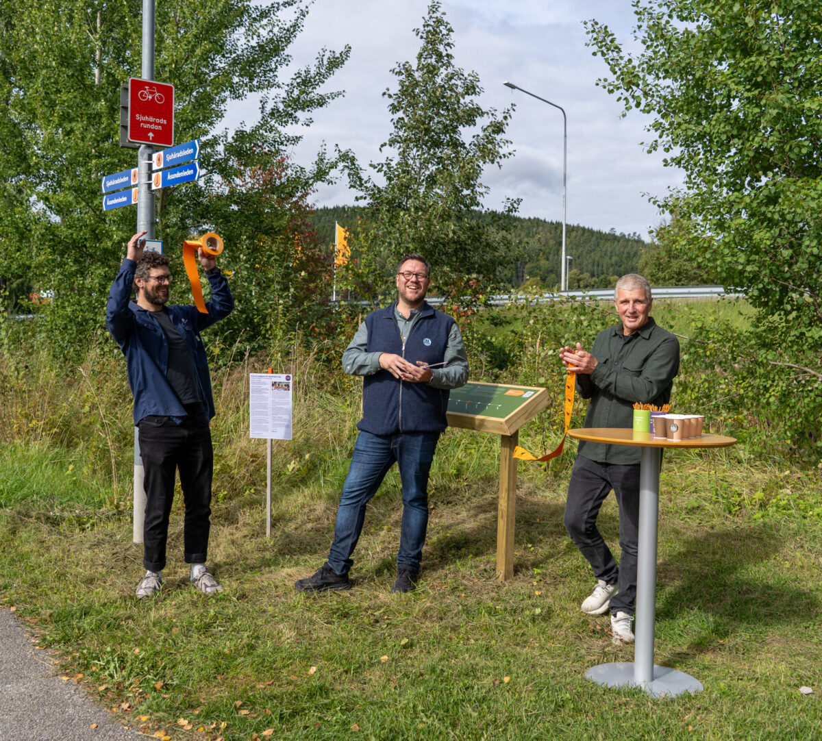 Tre glada personer på rad. Personen till vänster firar med händerna uppe, personen i mitten håller i en sax och personen till höger håller i en bit orange band. Foto.
