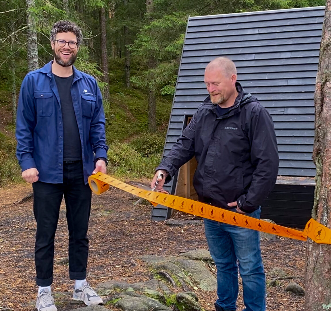 Person håller i ett orange band och en annan person klipper i det. I bakgrunden skymtar ett svart vindskydd och skog. Foto.