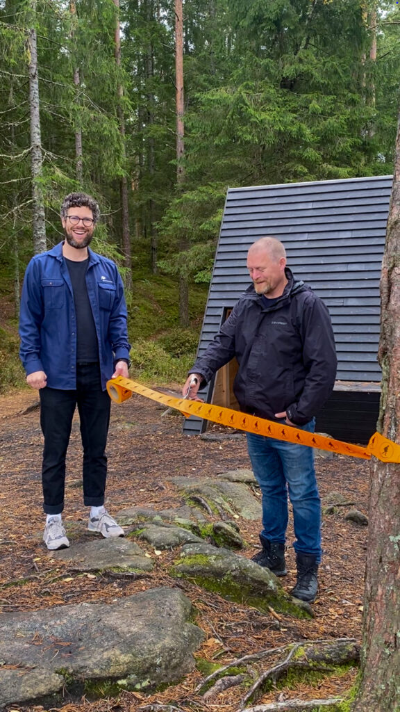 Person håller i ett orange band och en annan person klipper i det. I bakgrunden skymtar ett svart vindskydd och skog. Foto.